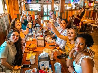 Smiling group of friends raising cups in a cheerful toast around a long wooden table during a casual brunch in a cozy rustic restaurant, plates, cups and condiments visible.