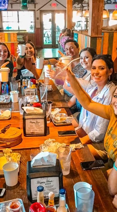 Smiling group of friends raising cups in a cheerful toast around a long wooden table during a casual brunch in a cozy rustic restaurant, plates, cups and condiments visible.