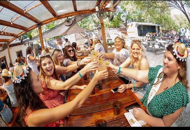Friends raising margarita glasses around a long wooden picnic table on a sunny outdoor patio, wearing mini sombreros and a sash for a festive bachelorette celebration.