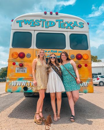 Three smiling women in sundresses—one wearing an "Engaged" sash and heart sunglasses—posing in front of a colorful yellow Texas party bus under a bright blue sky
