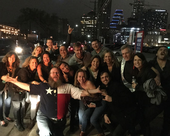 Lively group of friends posing for a nighttime downtown photo on a city street with lit skyscrapers and string lights; man in front wearing a Texas-flag shirt spreads his arms.