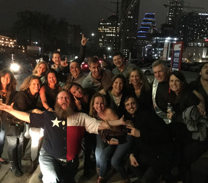 Lively group of friends posing for a nighttime downtown photo on a city street with lit skyscrapers and string lights; man in front wearing a Texas-flag shirt spreads his arms.
