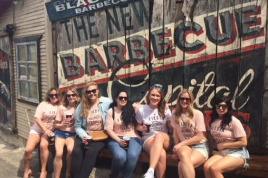Seven friends in matching Fiesta t-shirts holding drinks, sitting on a bench outside a street-side building with a large vintage wooden mural reading 'Barbecue'.