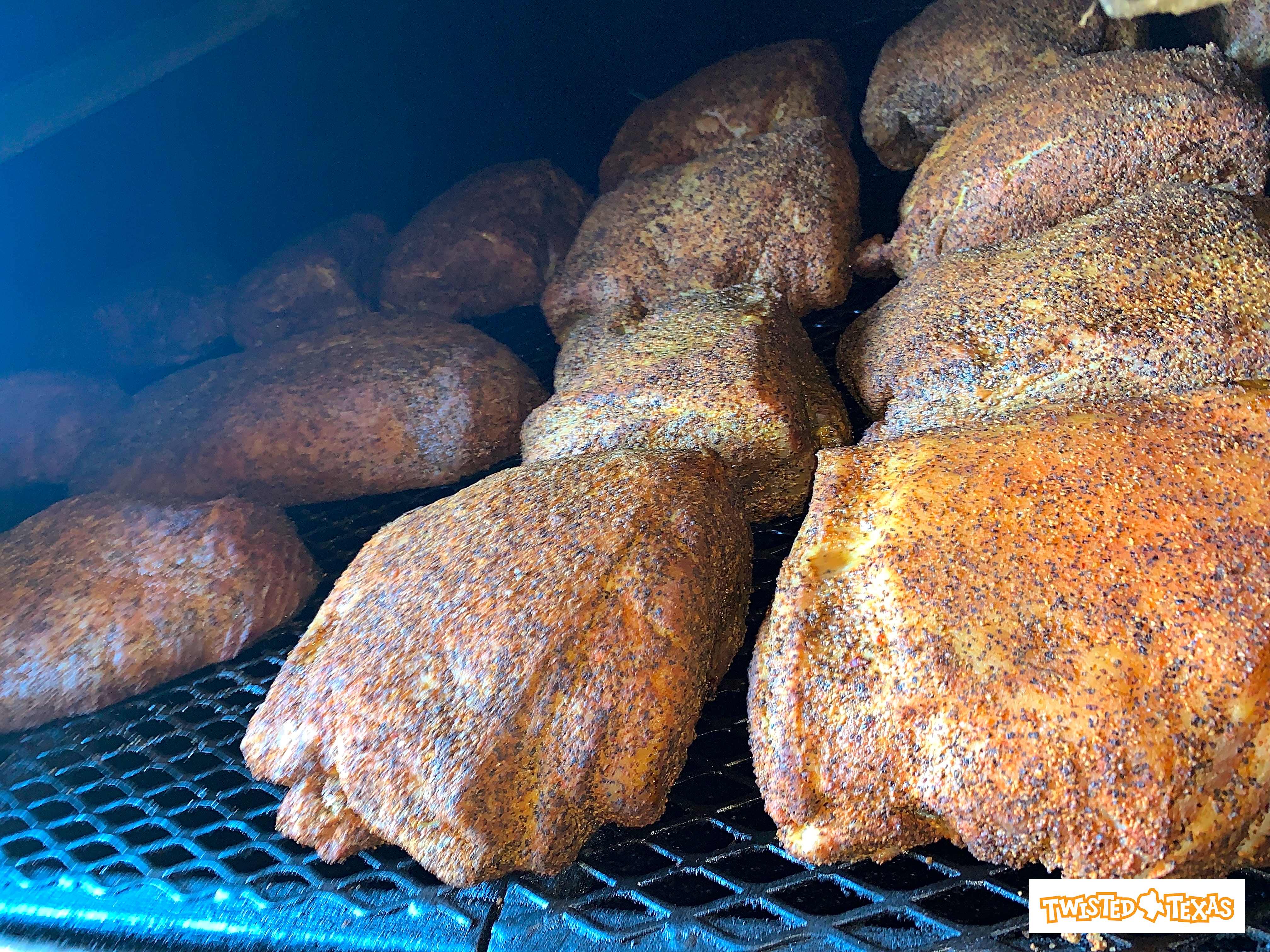 Multiple seasoned barbecue cuts with peppery crust smoking on metal racks inside a Texas-style smoker