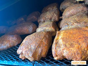 Multiple seasoned barbecue cuts with peppery crust smoking on metal racks inside a Texas-style smoker