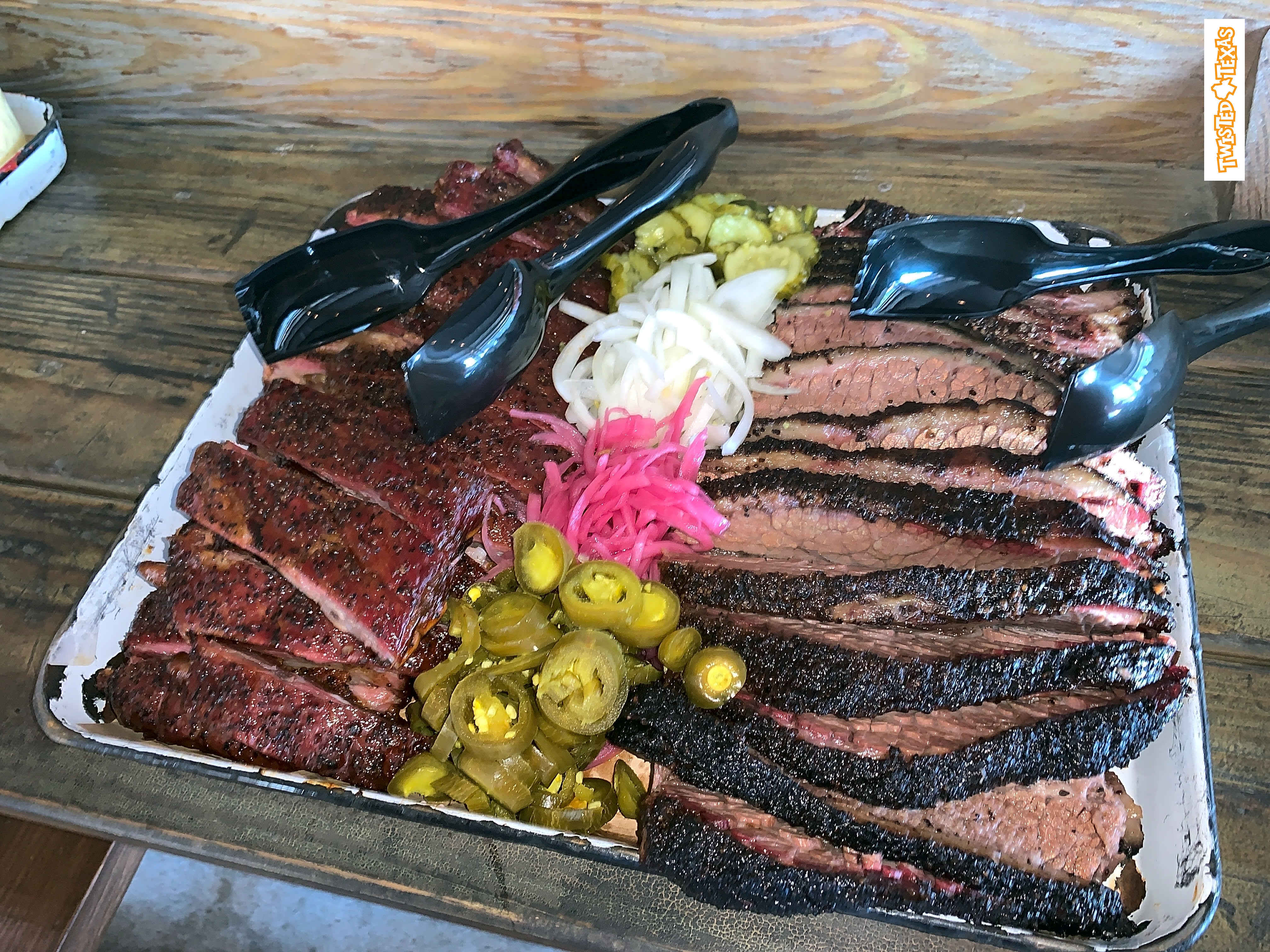 Heaping tray of sliced smoked brisket and ribs with pickled jalapeños, pickled onions and black serving tongs on a rustic wooden table — Texas-style barbecue platter.