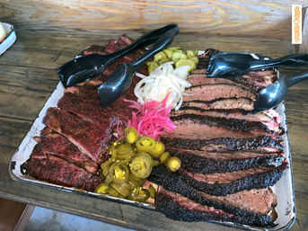 Heaping tray of sliced smoked brisket and ribs with pickled jalapeños, pickled onions and black serving tongs on a rustic wooden table — Texas-style barbecue platter.