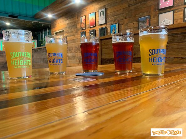 Flight of five craft beers in matching pint glasses on a polished wooden bar inside a rustic Texas taproom, from hazy pale ales to amber brews with framed artwork on the wood-paneled wall.