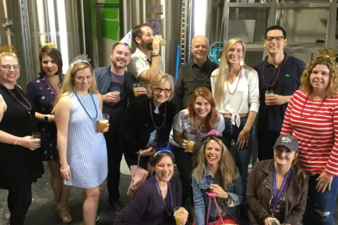 Smiling group of adults wearing party hats and beads, holding pints and posing in front of stainless-steel tanks at a craft brewery — casual brewery tour celebration.