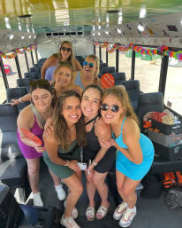 Seven friends smiling and posing inside a decorated party bus with colorful garlands, sunglasses, casual summer dresses, luggage and party props visible.