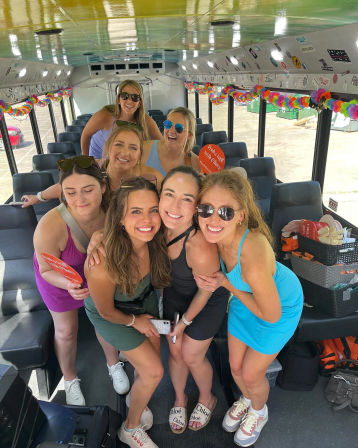 Seven friends smiling and posing inside a decorated party bus with colorful garlands, sunglasses, casual summer dresses, luggage and party props visible.