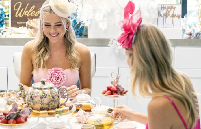Two people at a bright birthday tea party wearing decorative fascinators, sharing a floral teapot, macarons, fresh strawberries and tiered desserts.