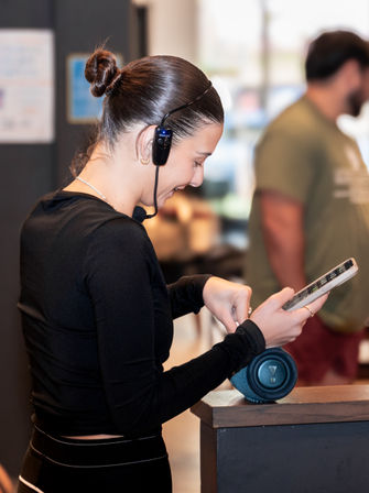Retail employee in black top wearing a headset and microphone, checking a tablet at a store counter with a portable Bluetooth speaker nearby