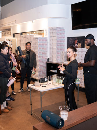 Retail pop-up product demo: woman presenting to a small group inside a shop with tall stacks of white boxes, demo table with glass jar and items, JBL speaker in foreground.