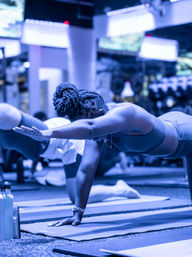 Participant in an indoor fitness studio performing a bird-dog plank on a yoga mat during a group class, showcasing balance, core strength, and an extended arm pose.
