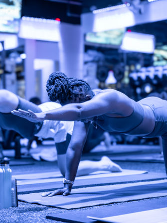 Participant in an indoor fitness studio performing a bird-dog plank on a yoga mat during a group class, showcasing balance, core strength, and an extended arm pose.