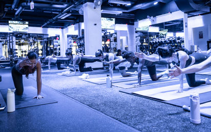 Group fitness class in a modern gym studio — participants on yoga mats practicing bird-dog core exercises with mirrors, weights and water bottles in the background.