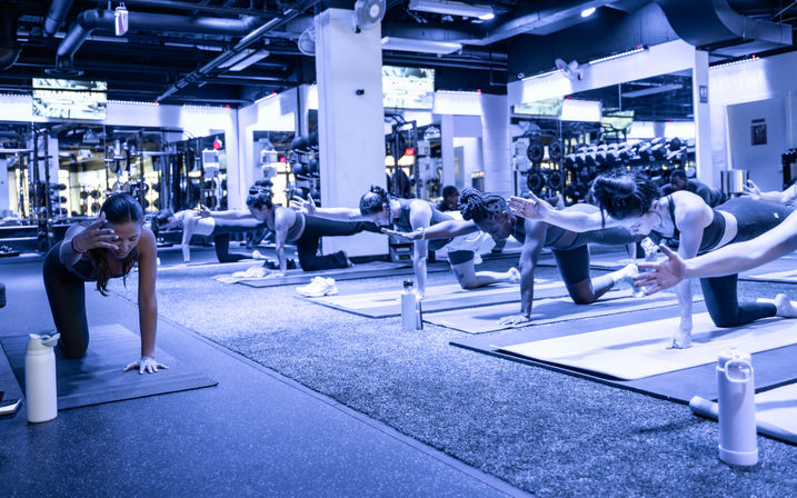 Group fitness class in a modern gym studio — participants on yoga mats practicing bird-dog core exercises with mirrors, weights and water bottles in the background.