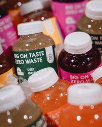Assorted colorful bottled juices with "Big on Taste, Zero Waste" labels chilling on ice in a market cooler