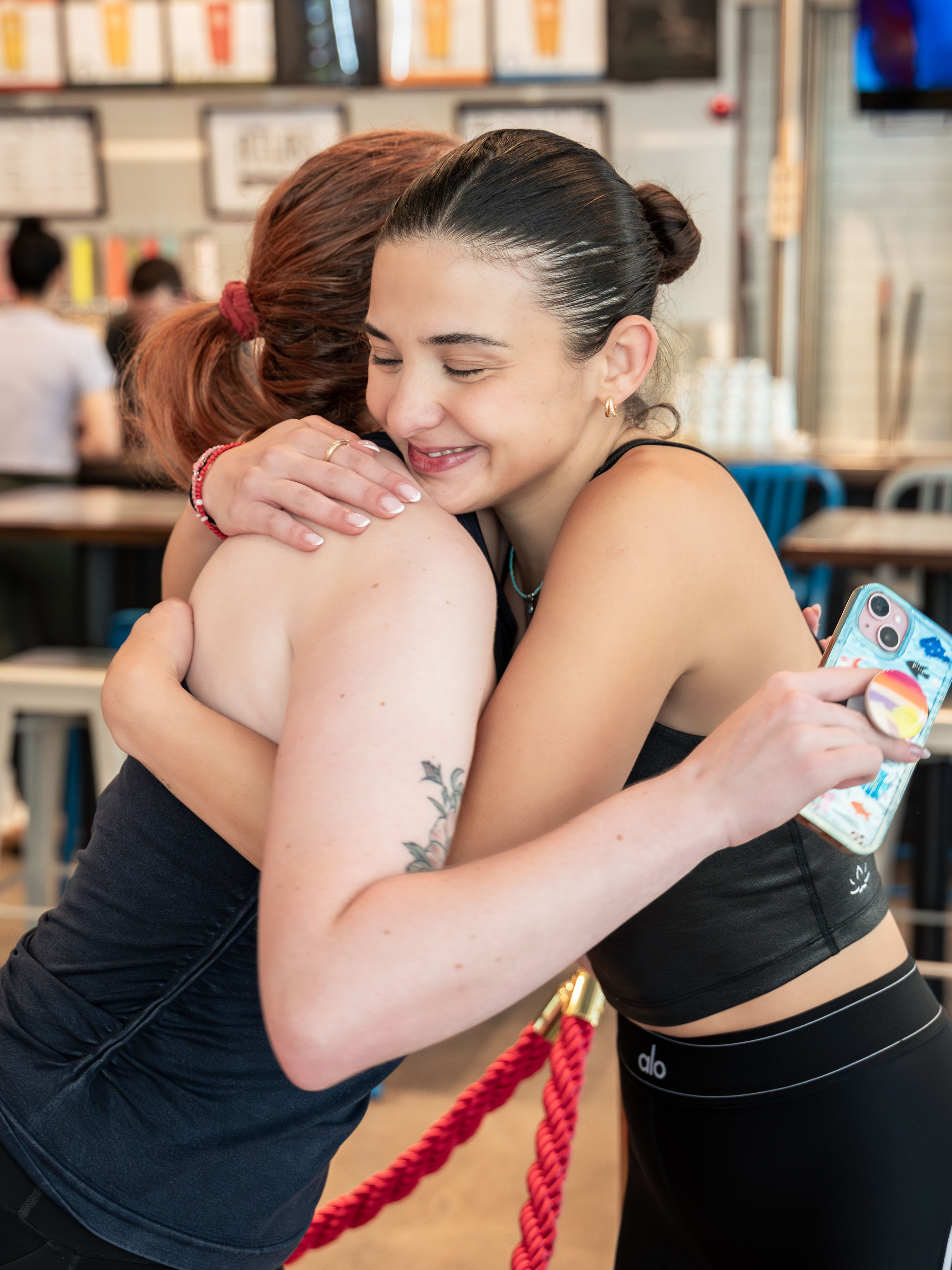 Smiling young women in athletic wear hug inside a bright cafe-style eatery, one holding a colorful phone case.
