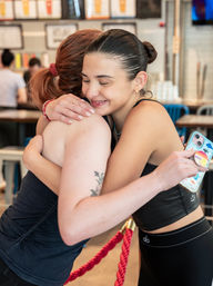 Smiling young women in athletic wear hug inside a bright cafe-style eatery, one holding a colorful phone case.