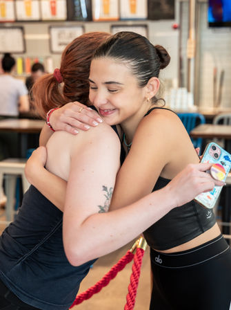 Smiling young women in athletic wear hug inside a bright cafe-style eatery, one holding a colorful phone case.