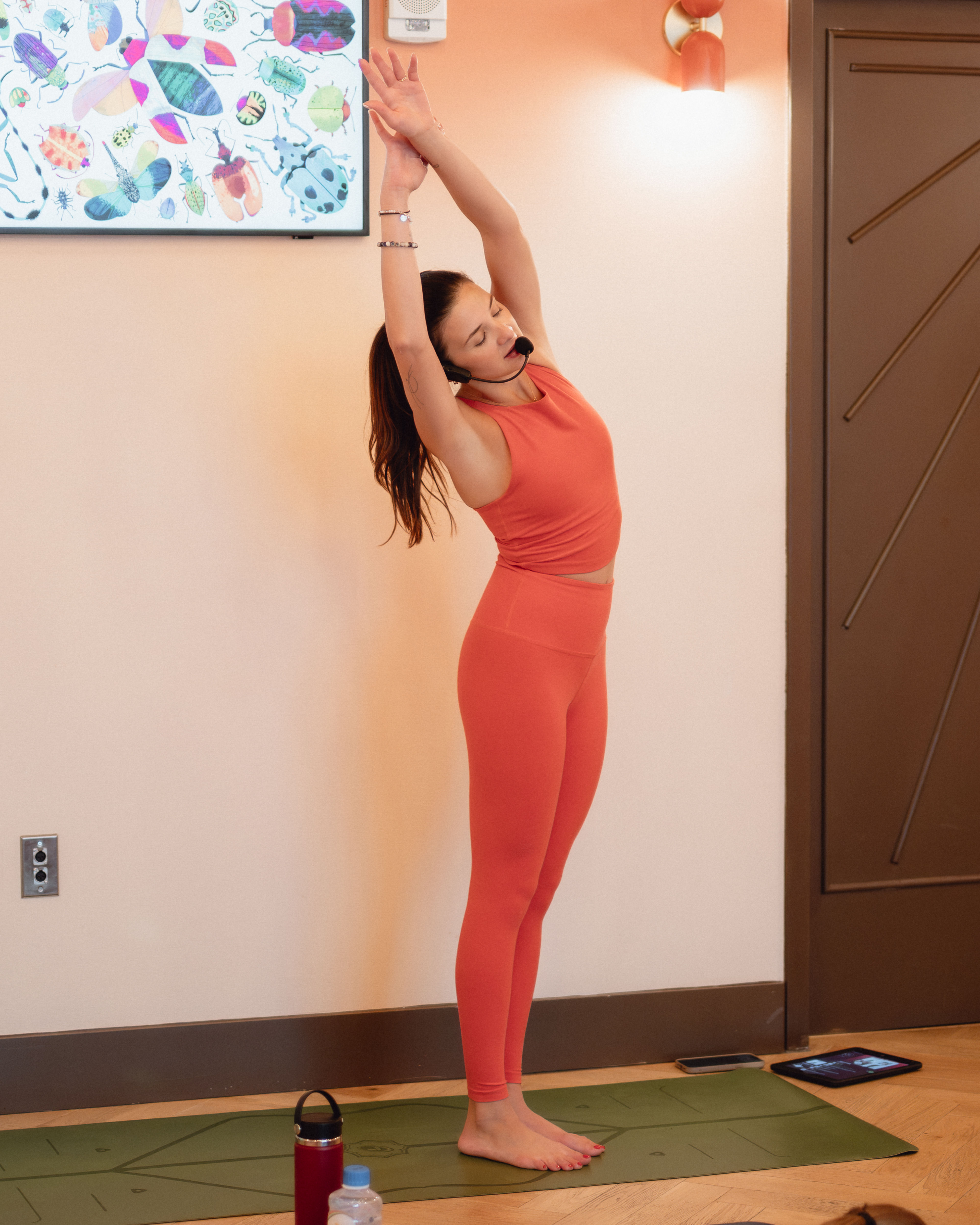 Indoor yoga instructor in coral activewear stretching arms overhead on a green mat, wearing a headset microphone with water bottle and tablet nearby and colorful wall art behind.