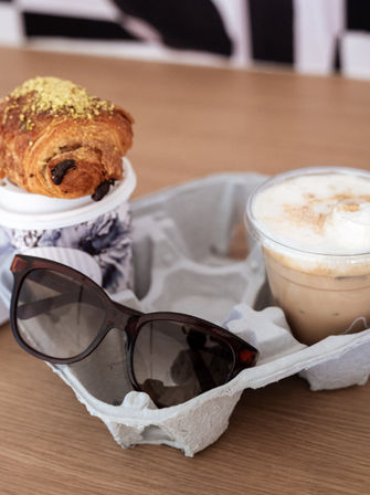 Sunglasses beside an iced latte and a chocolate-studded croissant topped with crushed pistachio in a cardboard cup carrier on a wooden café table