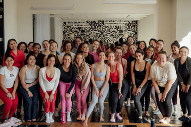 Smiling group of women in colorful activewear posing on yoga mats in a bright indoor fitness studio with hardwood floors, a mirrored wall and black-and-white mural — upbeat group yoga/fitness class vibe.