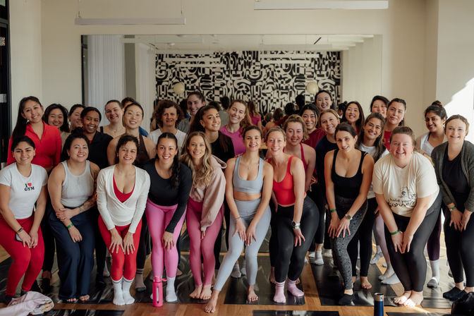 Smiling group of women in colorful activewear posing on yoga mats in a bright indoor fitness studio with hardwood floors, a mirrored wall and black-and-white mural — upbeat group yoga/fitness class vibe.