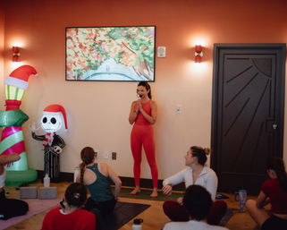 Indoor yoga studio group class: instructor in red activewear and headset leads students on mats, festive inflatable skeleton with Santa hat and colorful wall art in the background.