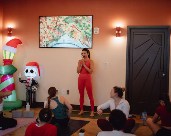 Indoor yoga studio group class: instructor in red activewear and headset leads students on mats, festive inflatable skeleton with Santa hat and colorful wall art in the background.
