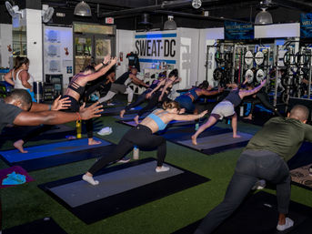 Energetic group workout in a Washington, D.C. indoor fitness studio — participants on mats lunging with arms extended, turf floor, weight racks and mirrors in the background.