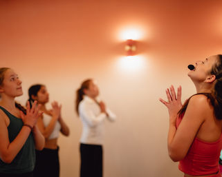 Enthusiastic instructor wearing a headset leads a warm-lit indoor group yoga class as young women stand with hands in prayer pose and eyes closed during a mindfulness session