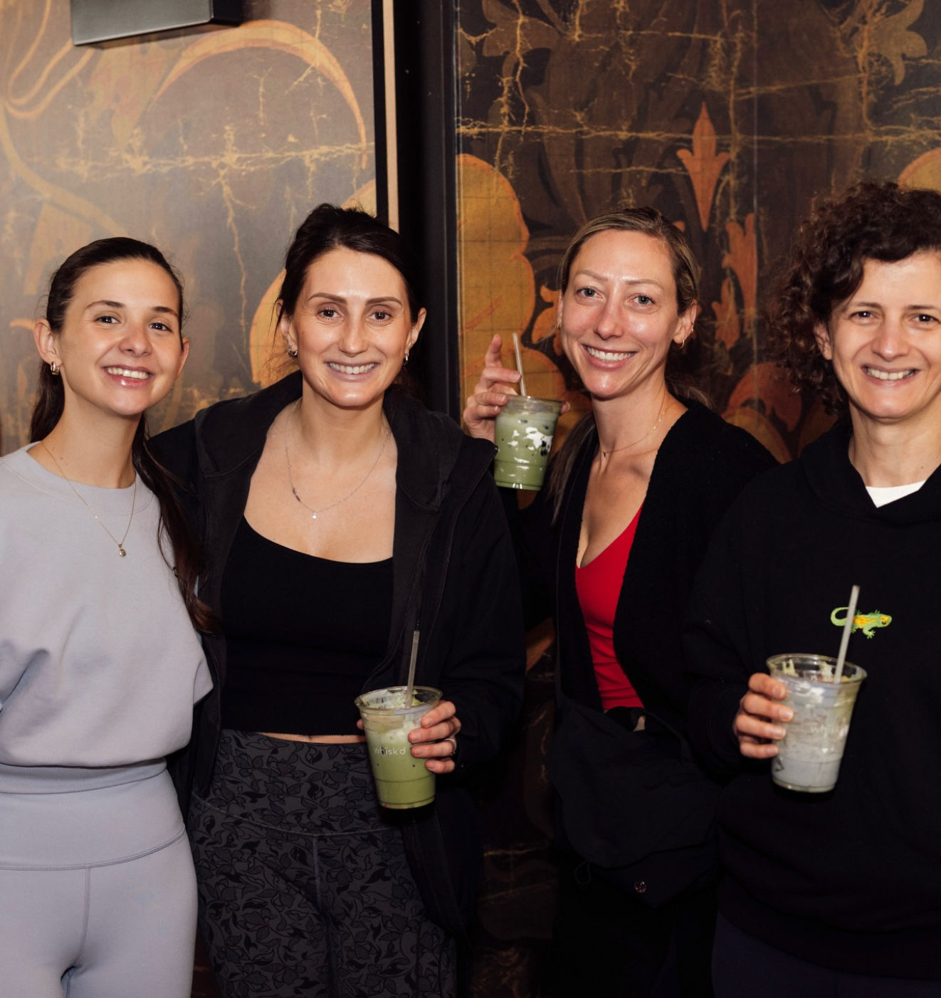 Four smiling women in casual activewear posing indoors in front of a decorative mural, each holding iced matcha drinks in clear cups — cheerful friends at a cozy cafe