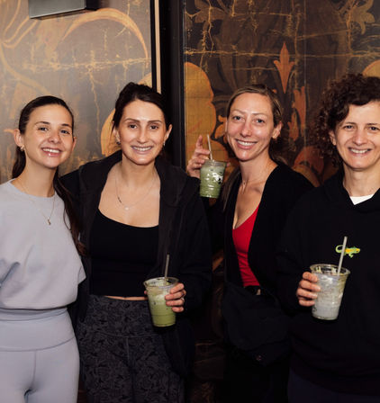 Four smiling women in casual activewear posing indoors in front of a decorative mural, each holding iced matcha drinks in clear cups — cheerful friends at a cozy cafe