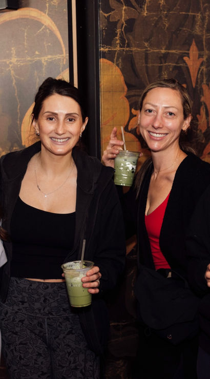 Four smiling women in casual activewear posing indoors in front of a decorative mural, each holding iced matcha drinks in clear cups — cheerful friends at a cozy cafe
