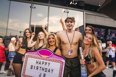 Group of smiling young adults celebrating a birthday on an outdoor bar patio — shirtless guest at center, friends holding sparklers, a marquee sign reading 'HAPPY BIRTHDAY JR' and a vodka bottle for the festivities.