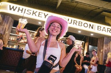 Smiling woman in a pink cowboy hat holding two plastic cocktails inside a lively whiskey-and-bourbon bar with a busy urban nightlife crowd in the background.