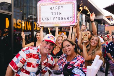 Energetic outdoor bar celebration with smiling crowd holding a ‘Happy 14th Anniversary’ marquee, sparklers, a bottle of vodka, and American-flag themed outfits.