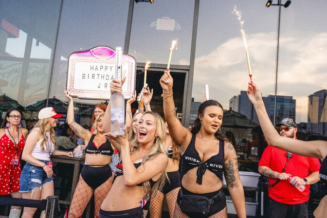 Cheerful group celebrating a birthday on an outdoor city patio at dusk, women in festive outfits holding sparklers and a large bottle beneath a light-up 'HAPPY BIRTHDAY' marquee with glass windows and skyline reflections.
