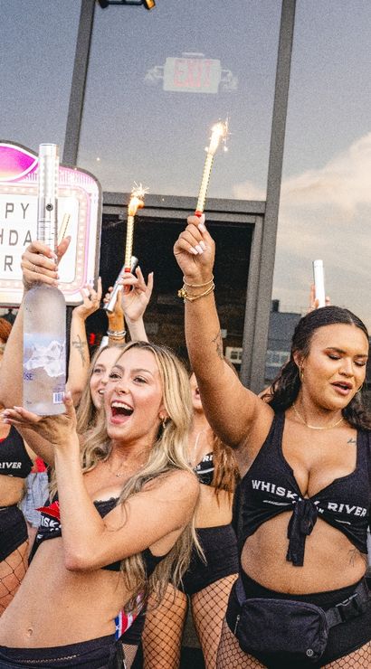 Cheerful group celebrating a birthday on an outdoor city patio at dusk, women in festive outfits holding sparklers and a large bottle beneath a light-up 'HAPPY BIRTHDAY' marquee with glass windows and skyline reflections.