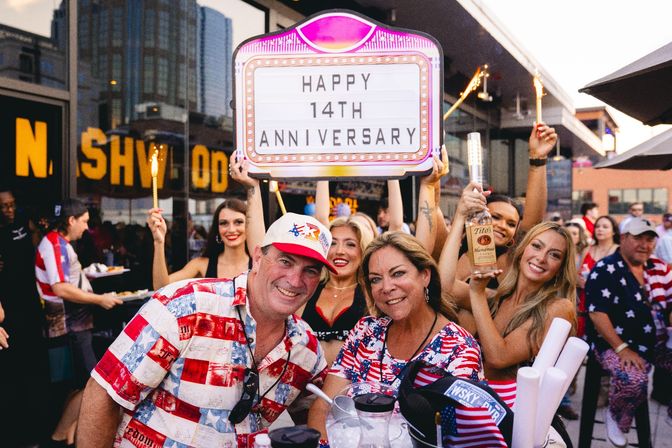 Lively downtown bar patio celebration with people in American-flag outfits holding sparklers, a bottle of vodka, and a marquee reading "Happy 14th Anniversary"