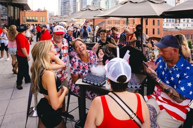 Smiling crowd at a rooftop patio party with downtown skyline in the background, guests wearing American-flag outfits and hats, chatting and enjoying drinks under umbrellas.
