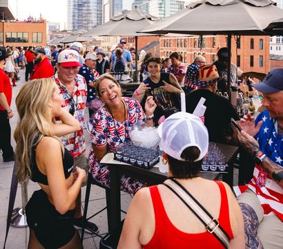 Smiling crowd at a rooftop patio party with downtown skyline in the background, guests wearing American-flag outfits and hats, chatting and enjoying drinks under umbrellas.