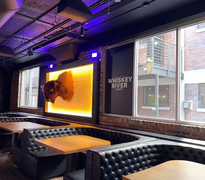 Industrial-style downtown bar interior with tufted black leather booths, wooden tables, exposed brick and ceiling pipes, large windows, and a glowing amber-backed mounted buffalo head display.