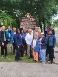 Group of adults smiling and posing by a historic school marker on a tree-lined neighborhood sidewalk