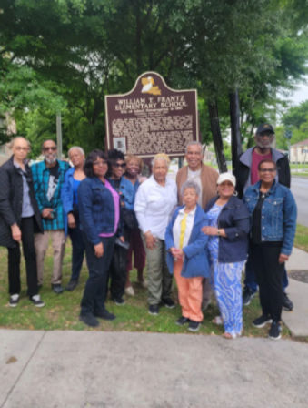 Group of adults smiling and posing by a historic school marker on a tree-lined neighborhood sidewalk