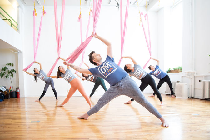 Group aerial yoga class in a bright white studio, participants holding pink silk hammocks while doing wide-legged side stretches on a polished hardwood floor