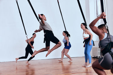 Adults in a bright indoor fitness studio doing an aerial suspension class — one person mid-jump while others laugh and practice moves on a hardwood floor.